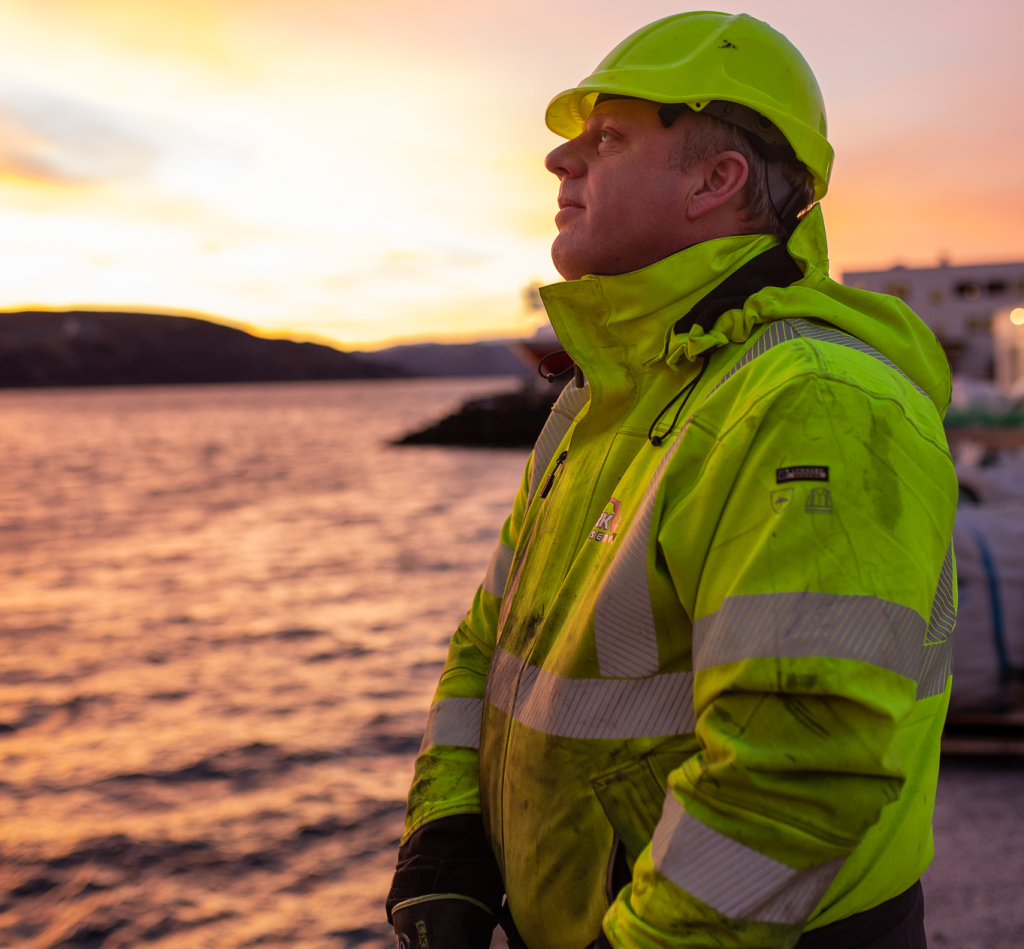 En arbeidskar iført en gul refleksjakke og hjelm står ved vannkanten og ser ut over havet i solnedgangen. Det varme lyset fra solen reflekteres i vannet og skaper en stemningsfull atmosfære. Jakken og hjelmen bærer logoen til "SRK Service", og mannen ser tankefull ut, kanskje etter en lang arbeidsdag.